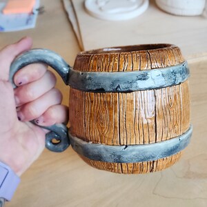 May include: A brown ceramic mug shaped like a barrel with a grey band around the top and bottom. The mug has a handle that is shaped like a twisted rope.