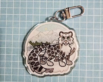 Op de afbeelding: Een heldere acryl sleutelhanger met een cartoon sneeuwluipaard. Het luipaard heeft een gevlekte vacht en staat tegen een besneeuwde bergachtergrond. De sleutelhanger heeft een zilveren sluiting. Het ontwerp bevat grijs, wit en groen.