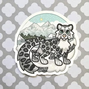 Puede incluir: Una pegatina con un leopardo de las nieves de dibujos animados con un pelaje manchado, en un paisaje montañoso nevado. El fondo incluye montañas, un cielo azul claro y un sol amarillo. La pegatina tiene un borde blanco.