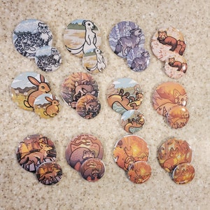 May include: A collection of round, illustrated animal buttons. The buttons feature various wildlife, including a snow leopard, rabbit, fox, and squirrel, in a variety of settings. Each button has a larger and smaller version. The color palette includes shades of brown, orange, and blue.