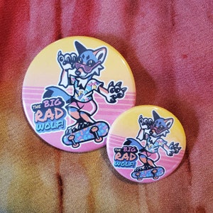 May include: Two round buttons with a cartoon wolf character wearing sunglasses and riding a skateboard. The buttons have a gradient background of yellow and pink with the text "THE BIG RAD WOLF!"