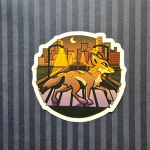 Könnte beinhalten: Ein Aufkleber mit einem stilisierten Kojoten, der einen Zebrastreifen vor einer Skyline bei Sonnenuntergang überquert. Der Kojote ist braun und hellbraun, und die Stadt ist in warmen Orange- und Gelbtönen beleuchtet. Ein Halbmond ist sichtbar.