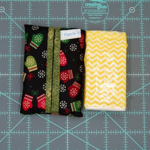 May include: A black fabric tissue holder with a pattern of red and green mittens and snowflakes. A label reads "Family T". Next to it is a rectangular tissue box with a yellow and white chevron pattern.