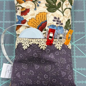 Puede incluir: Un coj&iacute;n de alfileres de tela floral morado con un borde de encaje y un bolsillo para guardar los materiales de costura. El bolsillo contiene un carrete de hilo blanco, una cinta m&eacute;trica y varios clips de colores. El coj&iacute;n de alfileres est&aacute; sobre una estera de corte gris.