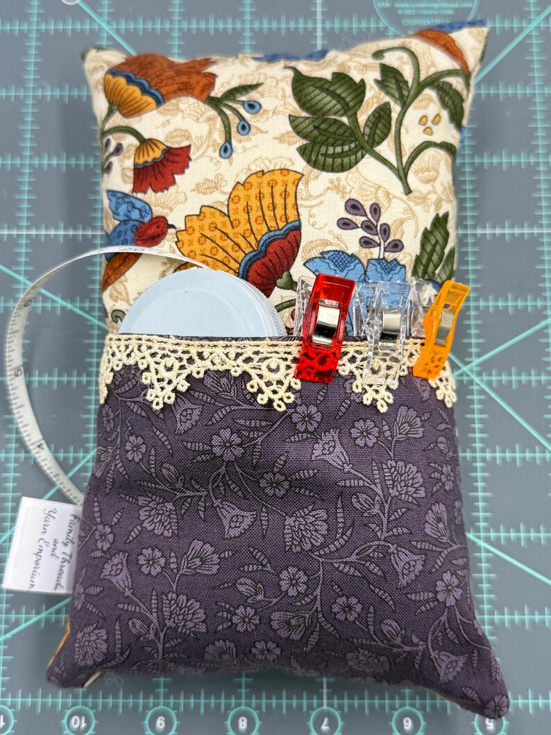 Puede incluir: Un coj&iacute;n de alfileres de tela floral morado con un borde de encaje y un bolsillo para guardar los materiales de costura. El bolsillo contiene un carrete de hilo blanco, una cinta m&eacute;trica y varios clips de colores. El coj&iacute;n de alfileres est&aacute; sobre una estera de corte gris.