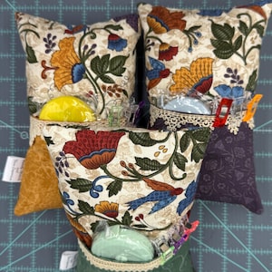 Puede incluir: Tres alfileteros de tela con estampado floral y ribetes de diferentes colores. Cada alfiletero tiene un peque&ntilde;o recipiente de pl&aacute;stico transparente con un bot&oacute;n de colores en su interior. Los alfileteros est&aacute;n sobre una estera de corte verde.