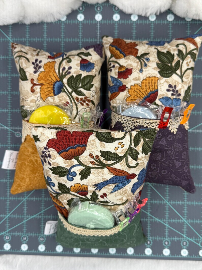 Puede incluir: Tres alfileteros de tela con estampado floral y ribetes de diferentes colores. Cada alfiletero tiene un peque&ntilde;o recipiente de pl&aacute;stico transparente con un bot&oacute;n de colores en su interior. Los alfileteros est&aacute;n sobre una estera de corte verde.