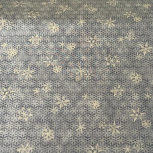 May include: A fabric with a repeating pattern of snowflakes in shades of white and cream against a gray background. The snowflakes are scattered across a geometric, diamond-shaped pattern.