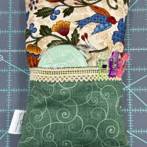 Puede incluir: Una almohada de tela verde con un patr&oacute;n de remolinos blancos. La almohada tiene un bolsillo con un borde de encaje y un forro de tela floral verde y blanco. El bolsillo contiene una herramienta de costura de pl&aacute;stico verde y clips de costura de colores.