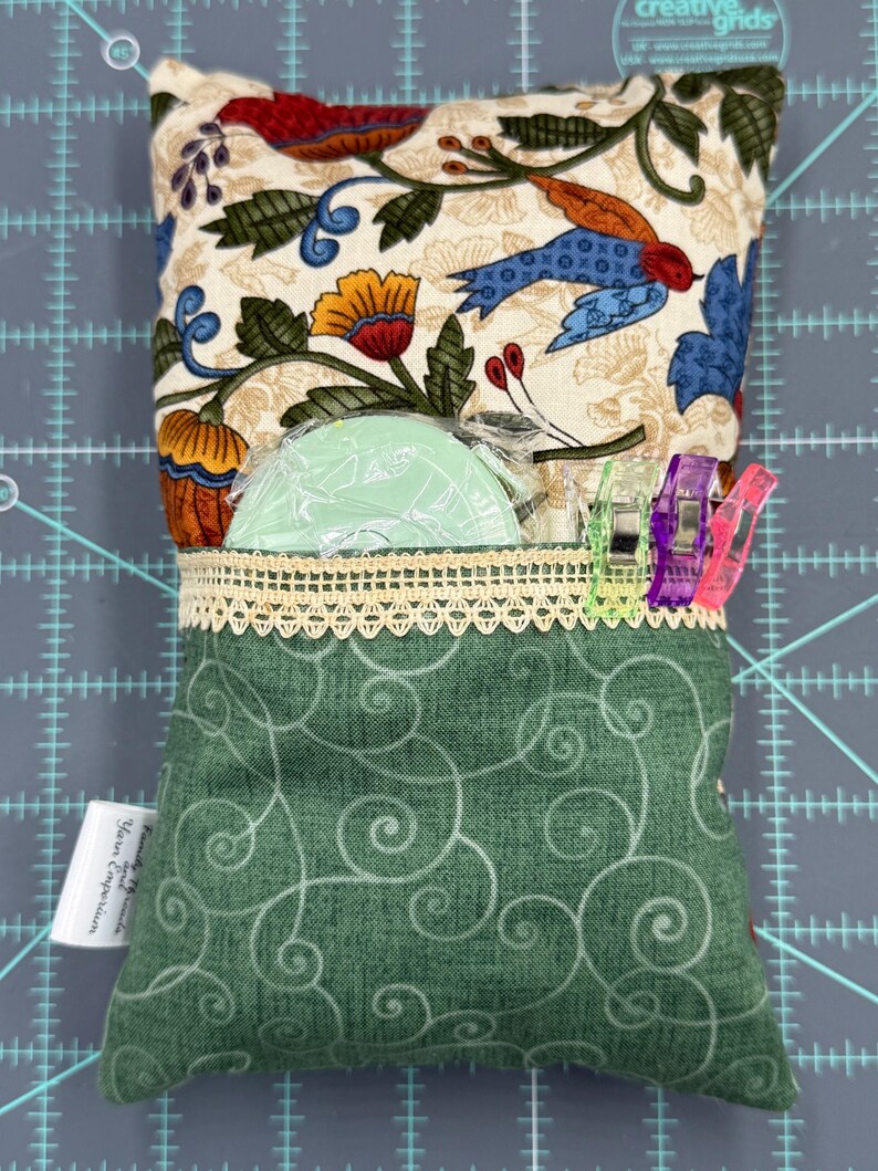 Puede incluir: Una almohada de tela verde con un patr&oacute;n de remolinos blancos. La almohada tiene un bolsillo con un borde de encaje y un forro de tela floral verde y blanco. El bolsillo contiene una herramienta de costura de pl&aacute;stico verde y clips de costura de colores.