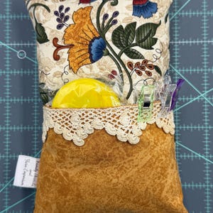 Puede incluir: Un coj&iacute;n de alfileres de tela marr&oacute;n con estampado floral y ribete de encaje. El coj&iacute;n est&aacute; relleno con un c&iacute;rculo de espuma amarillo y tiene una peque&ntilde;a etiqueta que dice "Family Heirlooms and Yara Emporium".