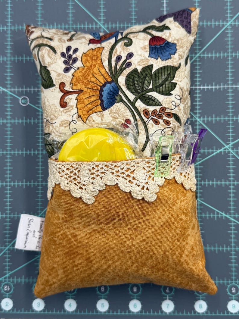Puede incluir: Un coj&iacute;n de alfileres de tela marr&oacute;n con estampado floral y ribete de encaje. El coj&iacute;n est&aacute; relleno con un c&iacute;rculo de espuma amarillo y tiene una peque&ntilde;a etiqueta que dice "Family Heirlooms and Yara Emporium".