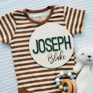 Puede incluir: Un body de beb&eacute; a rayas marrones y blancas con un letrero de madera que dice "JOSEPH Blake" en letras verdes y negras. Un oso de peluche blanco y un mordedor de madera con cuentas grises y blancas tambi&eacute;n est&aacute;n en la imagen.