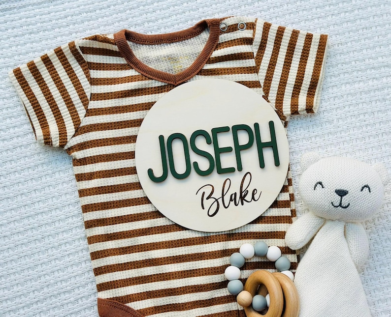 Puede incluir: Un body de beb&eacute; a rayas marrones y blancas con un letrero de madera que dice "JOSEPH Blake" en letras verdes y negras. Un oso de peluche blanco y un mordedor de madera con cuentas grises y blancas tambi&eacute;n est&aacute;n en la imagen.
