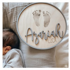 May include: A circular wooden plaque with a grey trim displays two baby footprints and the name "Azariah" in a cursive font, with "LUCA" beneath. The plaque is made of light-coloured wood.