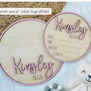 May include: Two round wooden baby announcement signs with pink trim. One sign has the name "Kinsley" in pink script. The other sign has the name "Kinsley" and lines for date, time, weight, and length. The text "NOVA" is also visible.