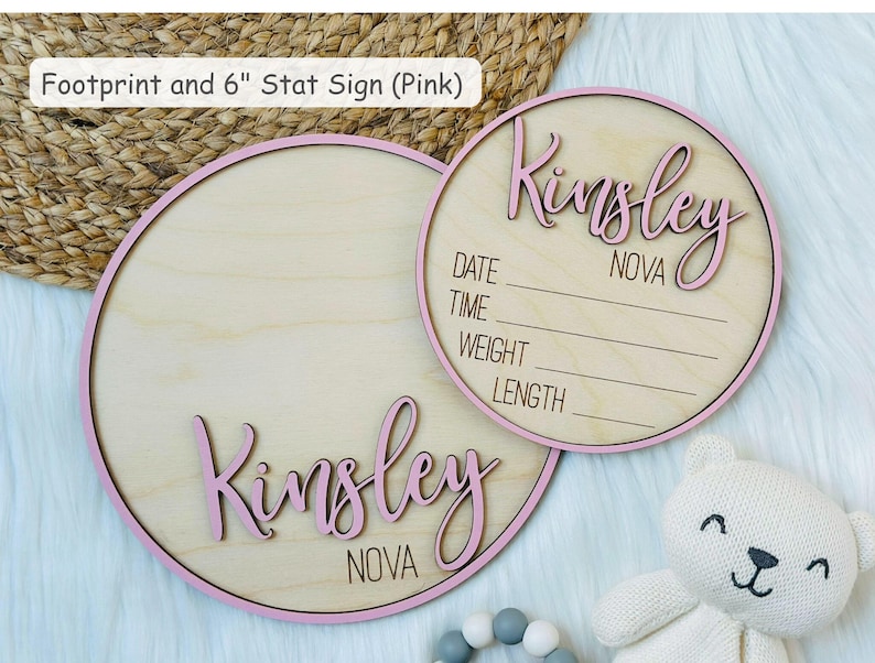 May include: Two round wooden baby announcement signs with pink trim. One sign has the name "Kinsley" in pink script. The other sign has the name "Kinsley" and lines for date, time, weight, and length. The text "NOVA" is also visible.