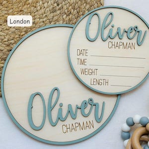 May include: Two round wooden baby announcement signs. The signs have a light wood colour with a teal border. One sign has the name "Oliver Chapman" in teal script. The other sign has the name "Oliver Chapman" and space to write the date, time, weight, and length.