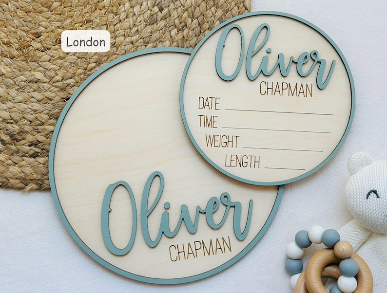 May include: Two round wooden baby announcement signs. The signs have a light wood colour with a teal border. One sign has the name "Oliver Chapman" in teal script. The other sign has the name "Oliver Chapman" and space to write the date, time, weight, and length.