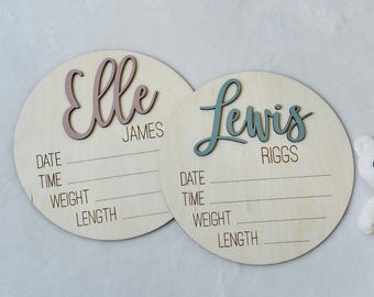 May include: Two circular light wood plaques featuring laser-engraved text for birth details including date, time, weight, and length. One plaque displays the name Elle James in raised mauve script, and the other shows Lewis Riggs in raised blue script.