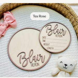May include: Two round wooden baby announcement signs with the name "Blair Heron" in a cursive font. One sign includes spaces for date, time, weight, and length. A pink bow and a white teddy bear are also in the image. The signs are "Tea Rose" coloured.