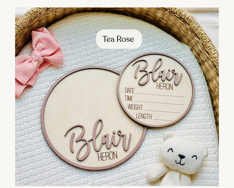 May include: Two round wooden baby announcement signs with the name "Blair Heron" in a cursive font. One sign includes spaces for date, time, weight, and length. A pink bow and a white teddy bear are also in the image. The signs are "Tea Rose" coloured.