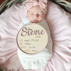 May include: A newborn baby wrapped in a white blanket, wearing a pink bow headband, and lying on a pink blanket. A wooden sign with the name "Stevie" and birth details: date, time, weight (709 g), and length (50.5 cm) is in the foreground.