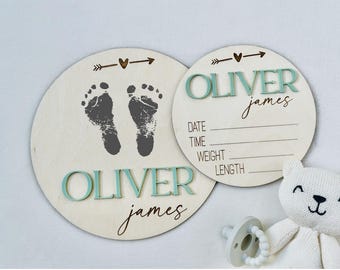 May include: Two circular wooden plaques featuring the name OLIVER james in raised mint green and black script lettering. One plaque displays dark gray baby footprints and an arrow with a heart. The other includes lines for date, time, weight, and length.