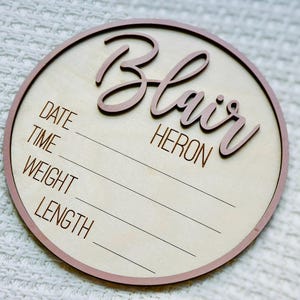 May include: A circular wooden birth announcement plaque with a light wood centre and a pink border. The name "Blair Heron" is displayed in a cursive font. The sign includes spaces to record the date, time, weight, and length.