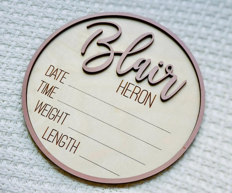 May include: A circular wooden birth announcement plaque with a light wood centre and a pink border. The name "Blair Heron" is displayed in a cursive font. The sign includes spaces to record the date, time, weight, and length.