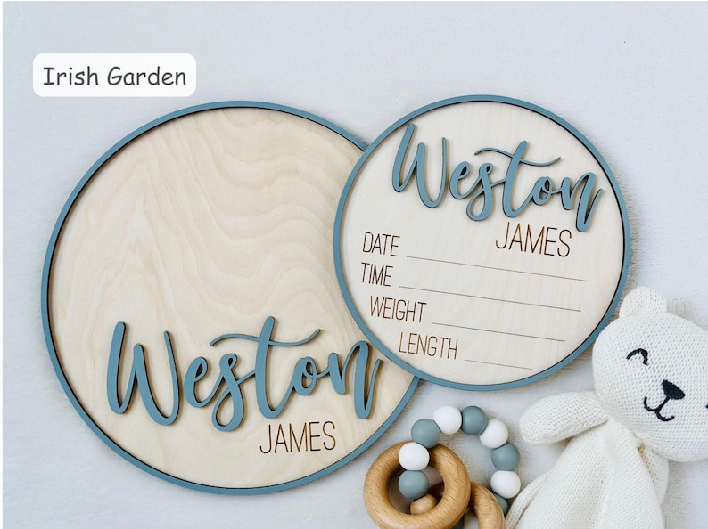 May include: Two wooden baby announcement signs with the name "Weston James" in a blue script font. One sign is a solid circle, the other has lines for date, time, weight, and length. A white teddy bear and a wooden teether are in the image.