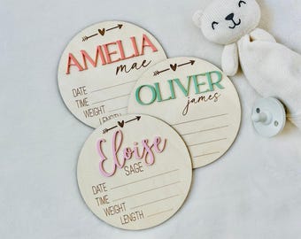 May include: Three circular wooden birth announcement signs featuring raised lettering in coral, teal, and pink. Each sign includes printed lines for date, time, weight, and length, with names Amelia Mae, Oliver James, and Eloise Sage displayed prominently.