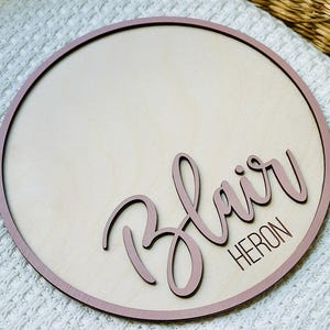 May include: A circular wooden sign with a light wood centre and a dusty rose border. The name "Blair" is written in cursive, and the surname "Heron" is below it, both in dusty rose. The sign rests on a white textured surface.