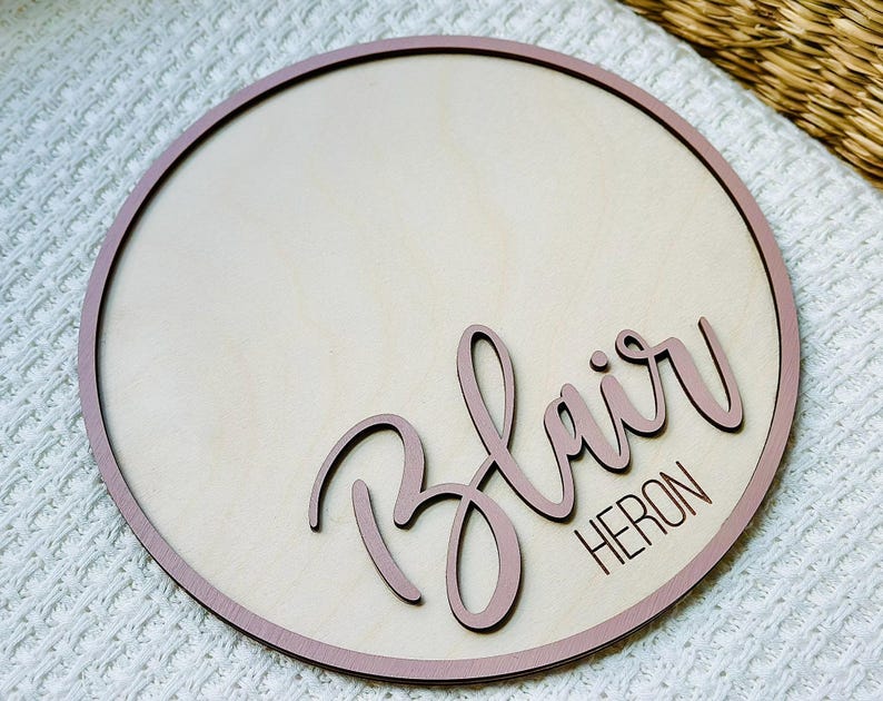 May include: A circular wooden sign with a light wood centre and a dusty rose border. The name "Blair" is written in cursive, and the surname "Heron" is below it, both in dusty rose. The sign rests on a white textured surface.