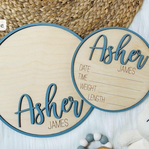 May include: Two wooden baby announcement signs. The signs are round with a blue border and the name "Asher James" in blue script. One sign has space for birth details. A white teddy bear and a wooden rattle are in the background.