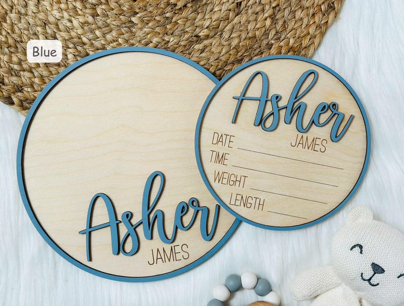 May include: Two wooden baby announcement signs. The signs are round with a blue border and the name "Asher James" in blue script. One sign has space for birth details. A white teddy bear and a wooden rattle are in the background.