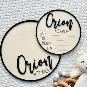 May include: Two circular wooden baby announcement plaques with black lettering and trim. The name "Orion Alexander" is displayed on both plaques. One plaque has space to write the date, time, weight, and length. A white teddy bear and wooden teething ring are in the image.