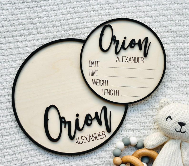 May include: Two circular wooden baby announcement plaques with black lettering and trim. The name "Orion Alexander" is displayed on both plaques. One plaque has space to write the date, time, weight, and length. A white teddy bear and wooden teething ring are in the image.