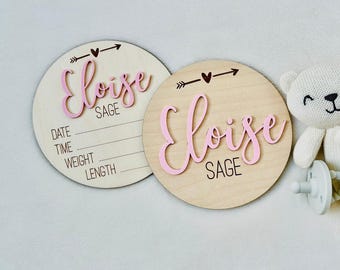 May include: Two circular light wood plaques featuring the name Eloise Sage in raised pink script lettering. One plaque includes engraved lines for date, time, weight, and length. Both feature a black arrow and heart graphic above the name.