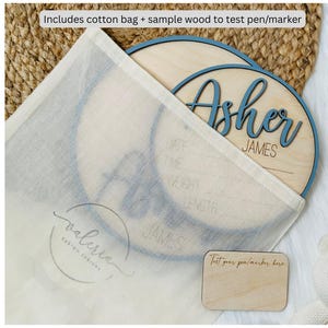 May include: A wooden baby name sign with the name "Asher" in blue script. The sign is inside a cotton bag, with a small wooden test piece. The text "Includes cotton bag + sample wood to test pen/marker" is visible.