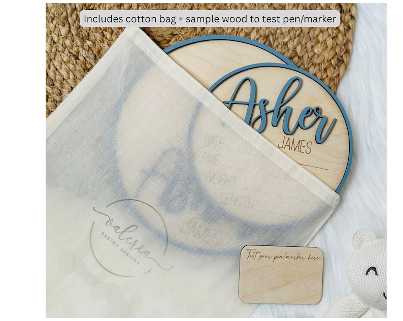 May include: A wooden baby name sign with the name "Asher" in blue script. The sign is inside a cotton bag, with a small wooden test piece. The text "Includes cotton bag + sample wood to test pen/marker" is visible.