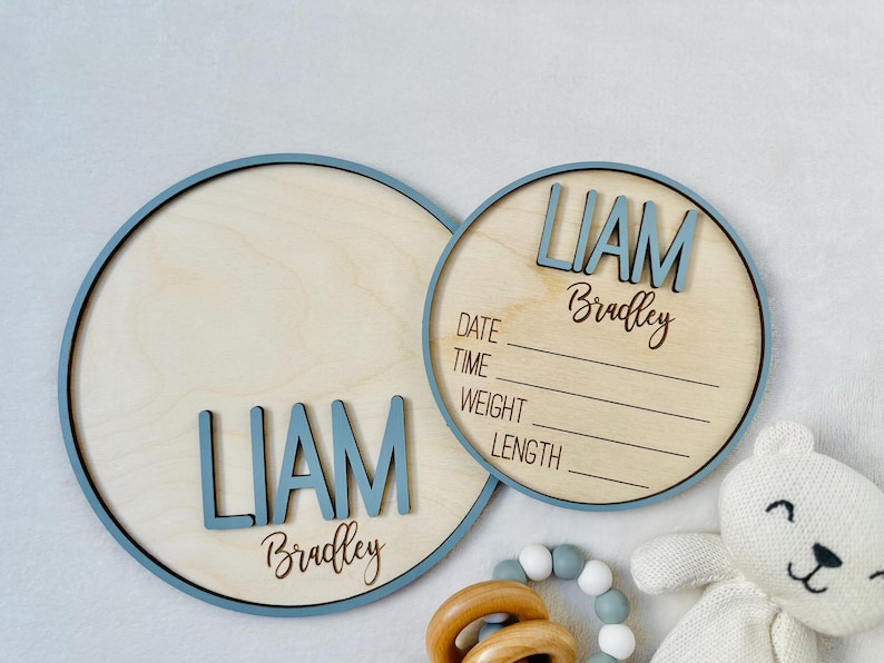 May include: Two wooden baby announcement signs. The larger sign is a circle with a blue border and the name "LIAM" in blue letters. The smaller sign has the name "LIAM Bradley" and lines for date, time, weight, and length. A white teddy bear is in the image.
