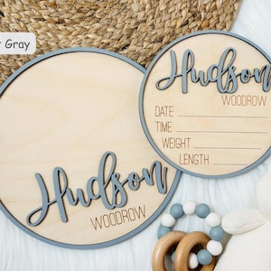May include: Two light wood round signs with light grey trim. One sign has the name "Hudson Woodrow" in script. The other sign has the name and space to write the date, time, weight, and length. A baby toy and stuffed bear are in the image.
