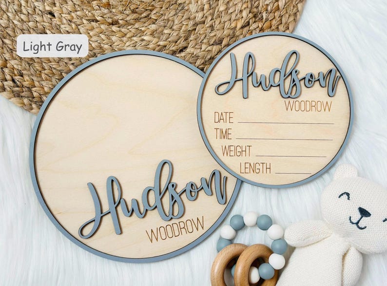 May include: Two light wood round signs with light grey trim. One sign has the name "Hudson Woodrow" in script. The other sign has the name and space to write the date, time, weight, and length. A baby toy and stuffed bear are in the image.