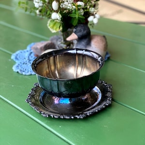 Puede incluir: Un cuenco de color plateado con un borde festoneado se encuentra sobre un plato decorativo a juego. El cuenco y el plato están sobre una superficie verde. Una figura de pato y un pequeño ramo de flores están en el fondo.