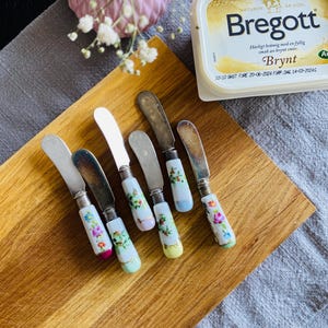 Puede incluir: Seis cuchillos de mantequilla con mangos con motivos florales sobre una tabla de cortar de madera. Los cuchillos tienen hojas plateadas y mangos blancos con diseños florales rosas, azules, amarillos y verdes.