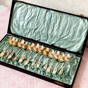 May include: A collection of silver spoons presented in a dark wooden box with a pale blue satin lining. The spoons are arranged in rows, reflecting light. The box is open, displaying the spoons and the satin interior.