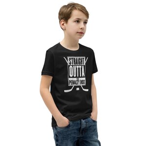 May include: Black youth t-shirt with a white graphic that says "Straight Outta The Penalty Box" with hockey sticks on either side of the text.