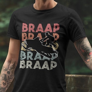 Pode incluir: Uma camiseta preta com um gráfico de estilo vintage de uma moto de neve e a palavra "BRAAP" repetida quatro vezes. O gráfico está em tons de vermelho, laranja, amarelo e azul.