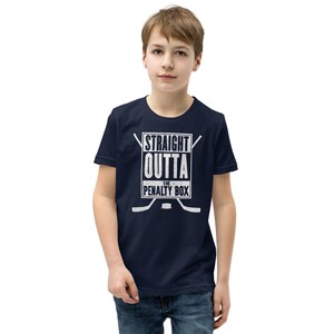 May include: A navy blue youth t-shirt with a white graphic that says "Straight Outta The Penalty Box" with two hockey sticks crossed behind the text.